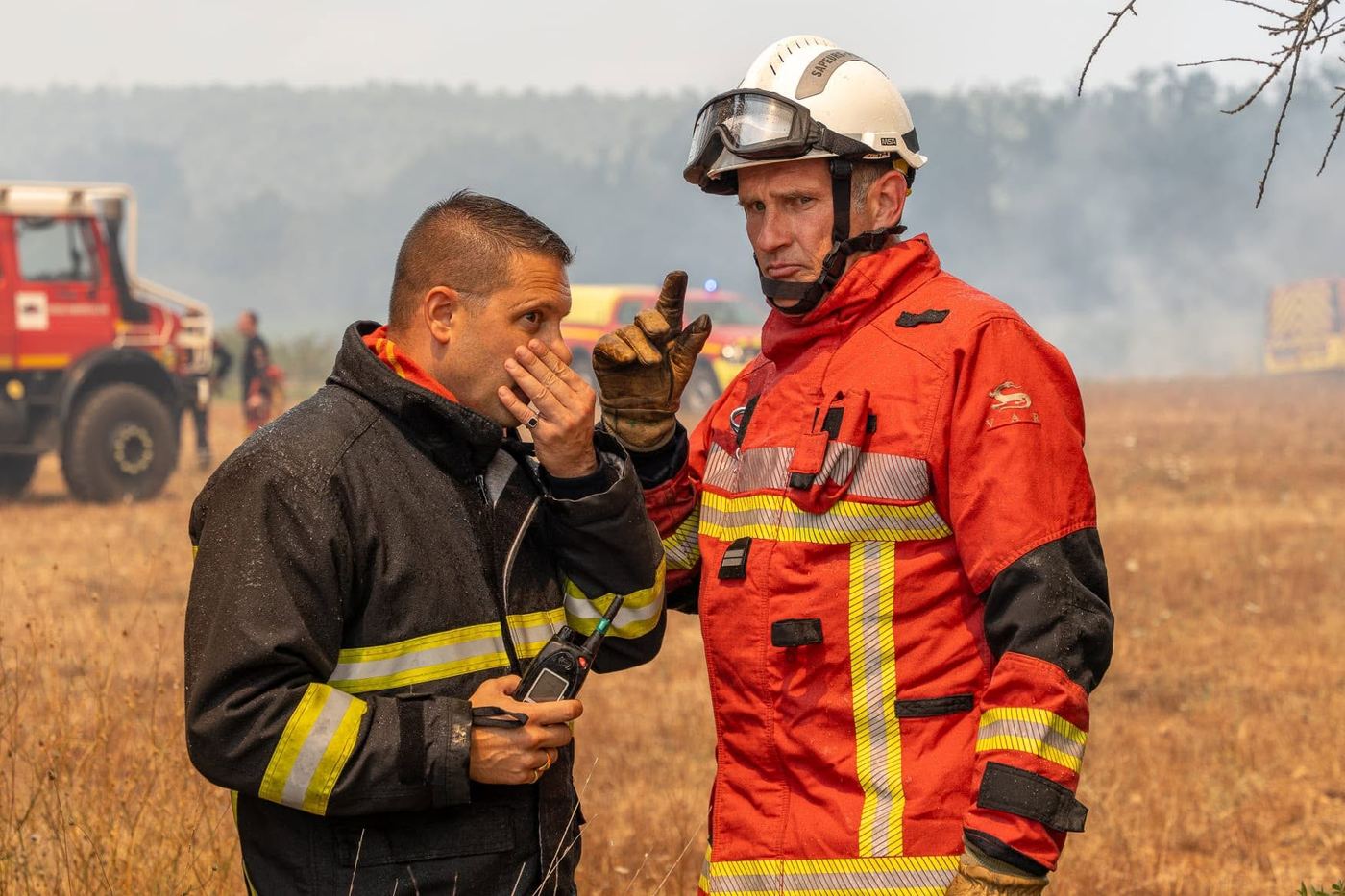 Twee brandweerlieden van de Sapeurs-Pompiers du Var overleggen aan de rand van het brandgebied in de Var, met op de achtergrond een tankautospuit en rook boven de heuvels.