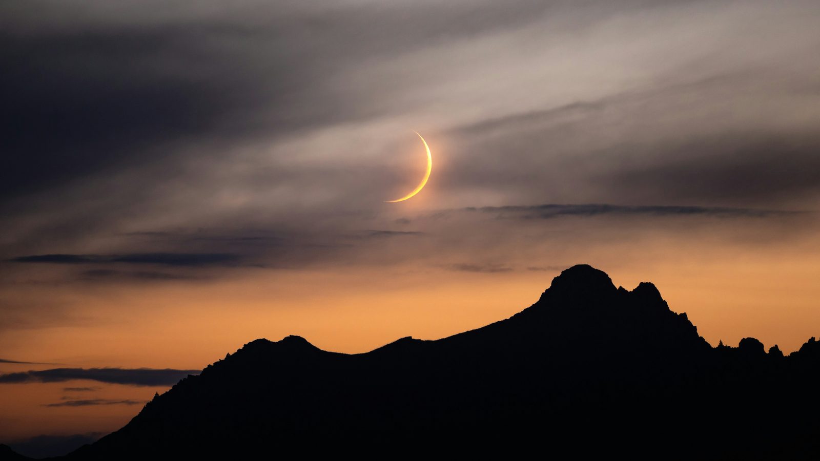 Smalle sikkel van een gedeeltelijk verduisterde zon, laag boven een donkere bergkam in een oranje avondlucht